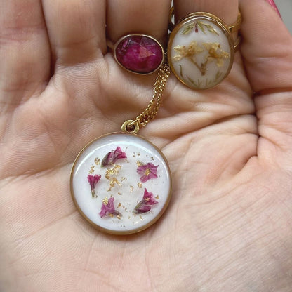Collar hecho a mano con flores naturales rosas y láminas doradas en resina de fondo blanco, sobre la palma de una mano